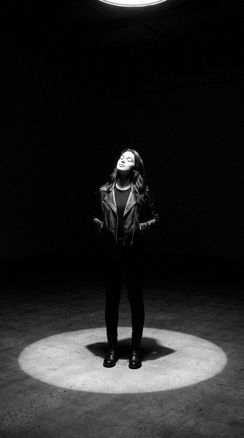 Nova Rai standing in a circle of light on a dark floor, looking up
