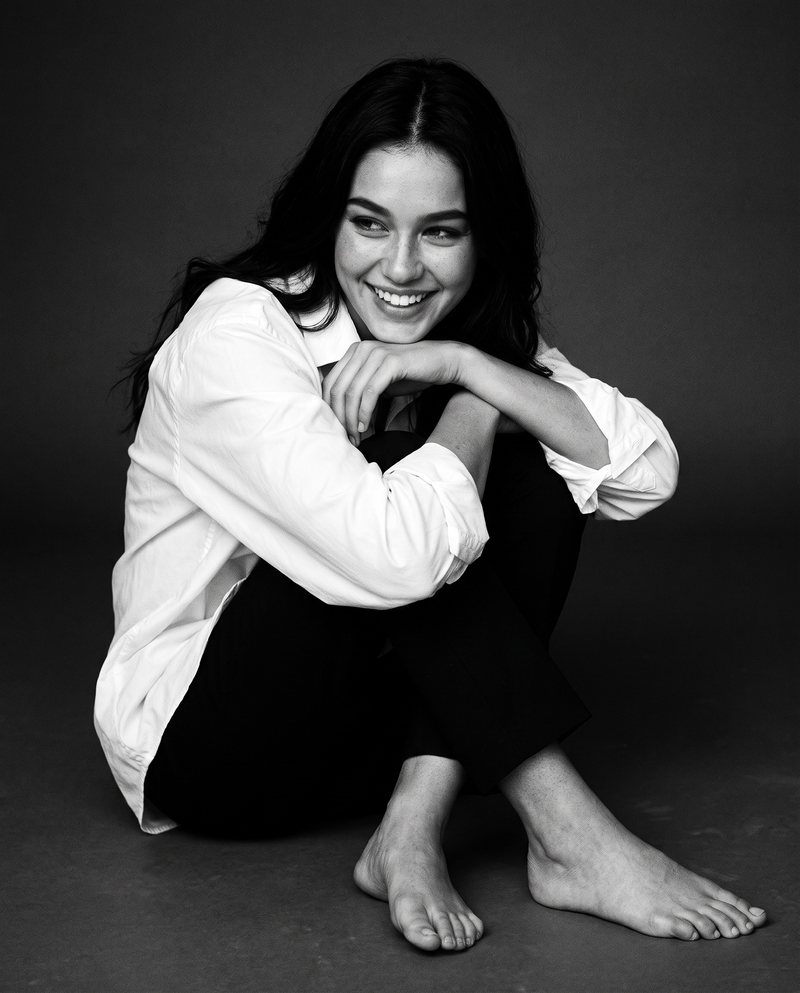 Nova Rai laughing on a studio floor with her arms around her knees, in black and white