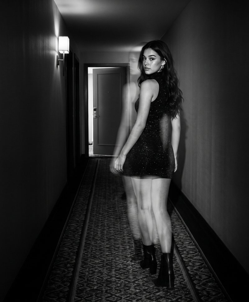 Nova Rai walking away down a hotel corridor and looking back, in black and white