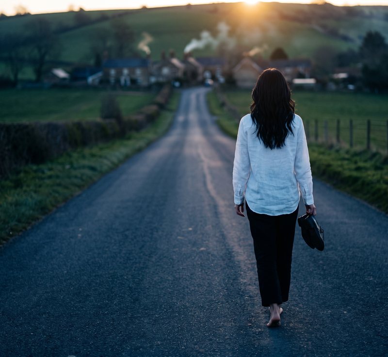 Nova Rai seen from behind, walking barefoot down a country road toward a village at sunrise, shoes in her hand