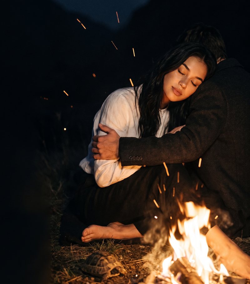 Nova Rai with her eyes closed, held close by a campfire as sparks rise