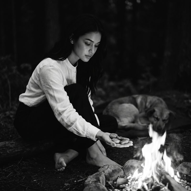Nova Rai by a campfire at night holding out a hand of small stones, a dog asleep nearby, in black and white