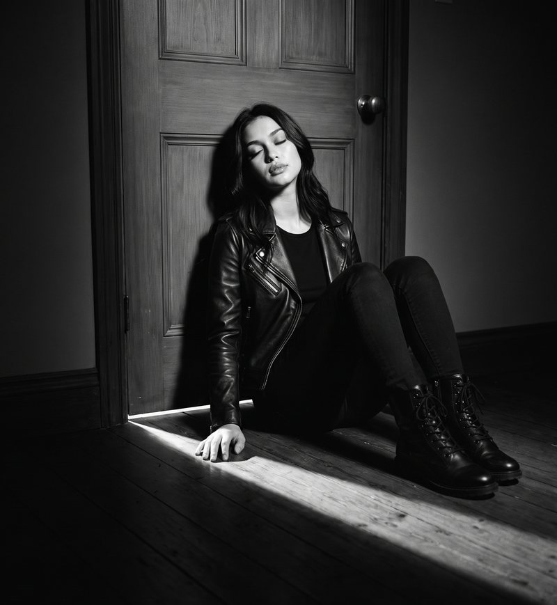 Nova Rai sitting on the floor by a door in a patch of light, in black and white