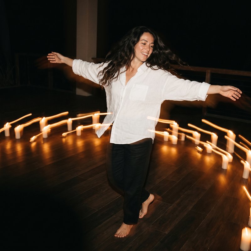 Nova Rai dancing barefoot on a wooden floor, arms wide and smiling, candles blurred into lines of light around her