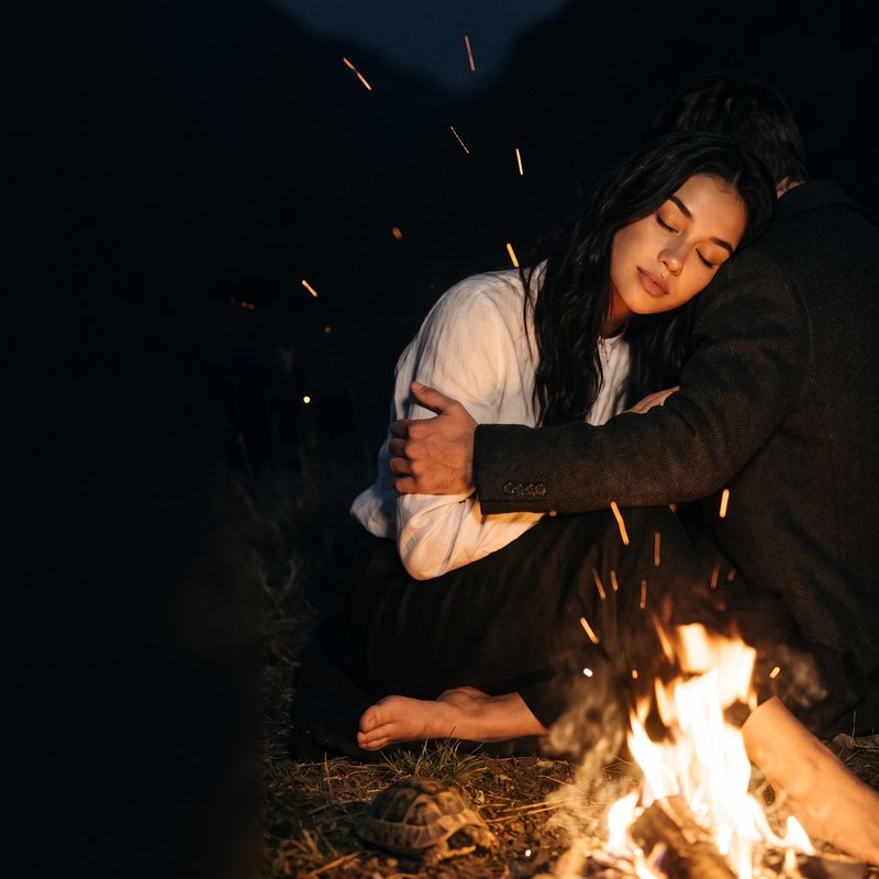 Nova Rai with her eyes closed, held by a man in a dark coat beside a campfire, sparks rising