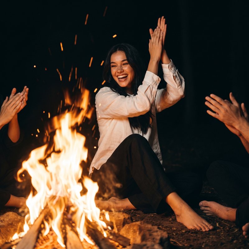 Nova Rai laughing and clapping by a bonfire at night, other hands clapping around her