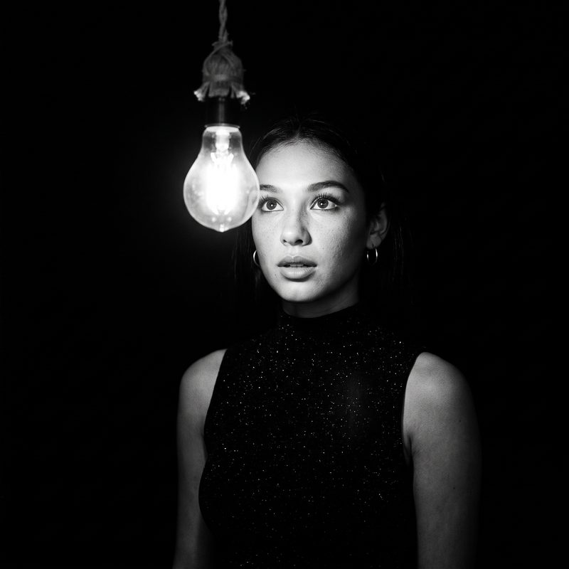 Nova Rai in black and white beside a single bare light bulb in the dark, looking past it